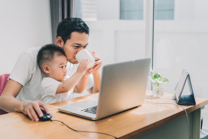 Chinese dad in front of a laptop, drinking a mug his infant son is helping to hold to him.