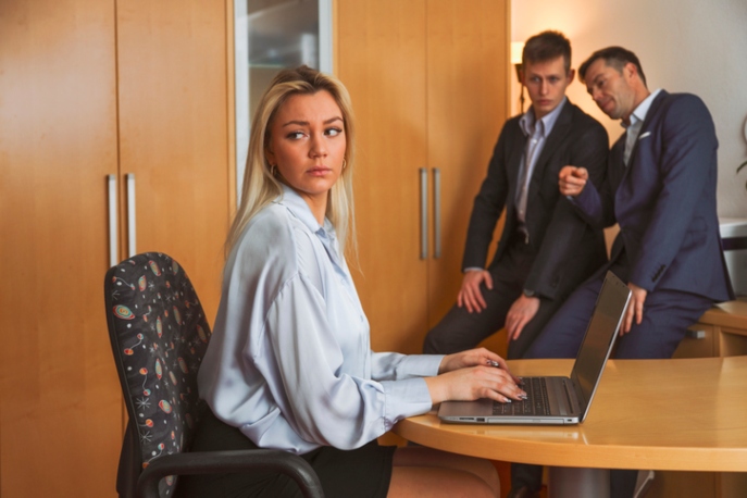 Woman in office looking dismayed while men visibly talk about her in the background.