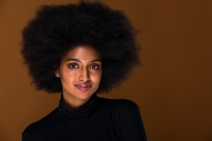 Black woman with afro in a black turtleneck and pink and yellow eyeshadow, smiling at the camera